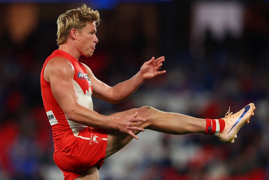 Side profile of a Sydney Swans AFL player with his leg raised to hip height in front of him, having just kicked a football.