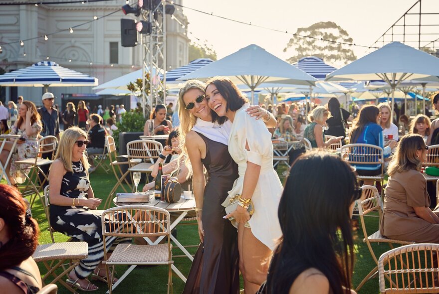 The Fashion Forecourt located outside the Royal Exhibition Building, with people socialising and enjoying a drink at tables  under shade umbrellas, and two friends laughing and standing close together as if posing for a photo.