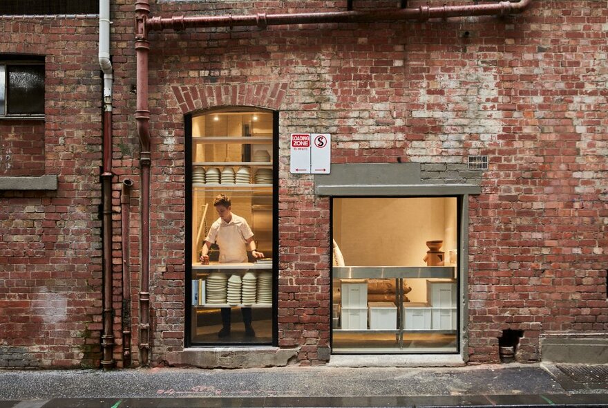 Exterior of an old brick building with windows and doors open to reveal a kitchen and a man in white in the window. 