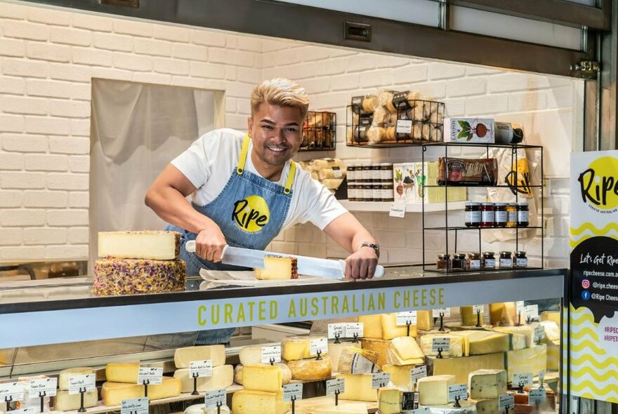 A smiling person slicing cheese with a very large knife, on the counter of a display cabinet that is filled with many different cheeses.