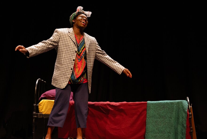 A woman on stage playing reknowned jazz singer and civil rights activist, Nina Simone, singing as she stands next to a simple bed.