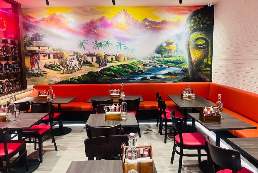 Brightly decorated restaurant interior, with a large mural on the rear wall, small square black tables and red cushioned chairs, and banquette seating all around the walls.