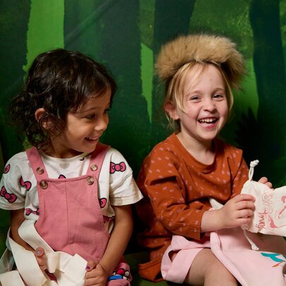 Two young children smiling while participating in an interactive play or educational activity against a green forest-themed backdrop, at Melbourne Museum.