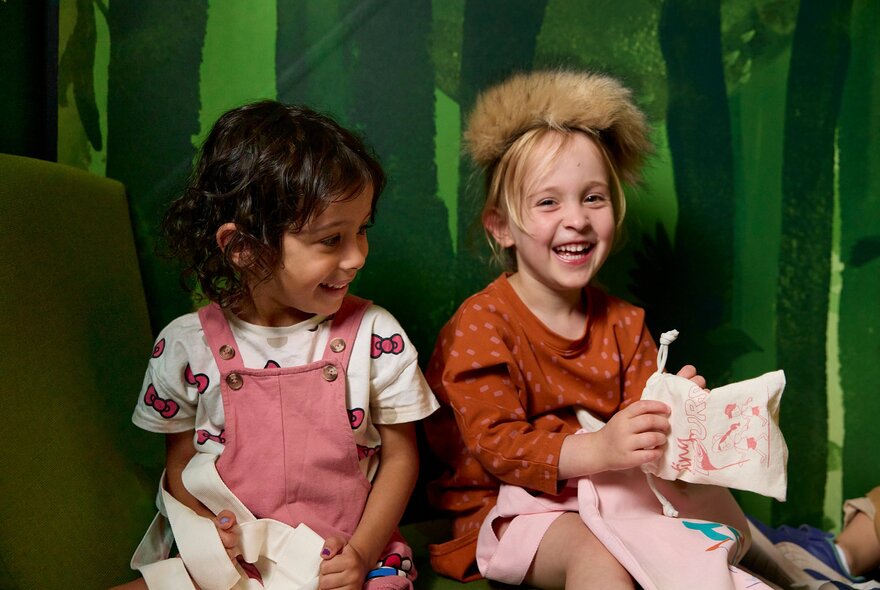 Two young children smiling while participating in an interactive play or educational activity against a green forest-themed backdrop, at Melbourne Museum.