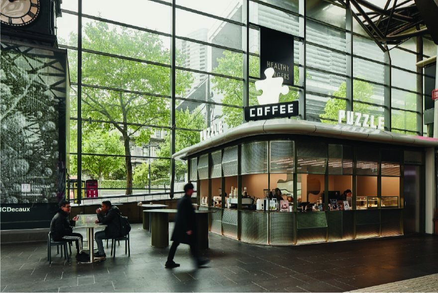 The Puzzle Coffee outlet at Southern Cross Station, with floor to ceiling glass windows letting in much natural light behind it, and people seated at a small table outside the cafe.