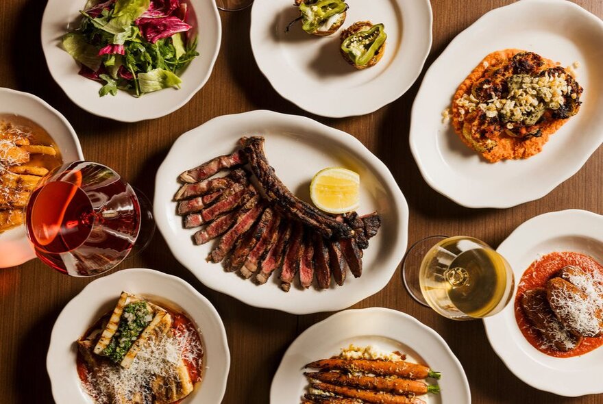An overhead shot of a wooden table spread with various dishes, including a central platter of sliced steak with a lemon wedge, and smaller plates of salads, roasted vegetables, and glasses of red and white wine.