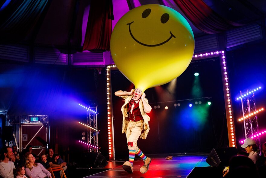A circus performer wearing a giant yellow smiley-face balloon as a hat, on stage at a children's show.