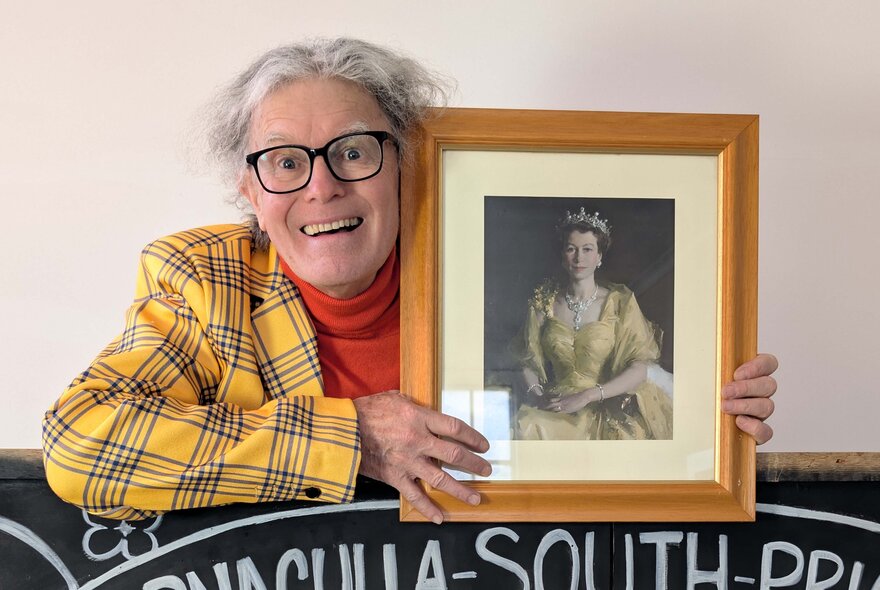 Comedian wearing yellow tartan jacket and red skivvy, with grey hair and spectacles, holding Wattle portrait of the Queen.