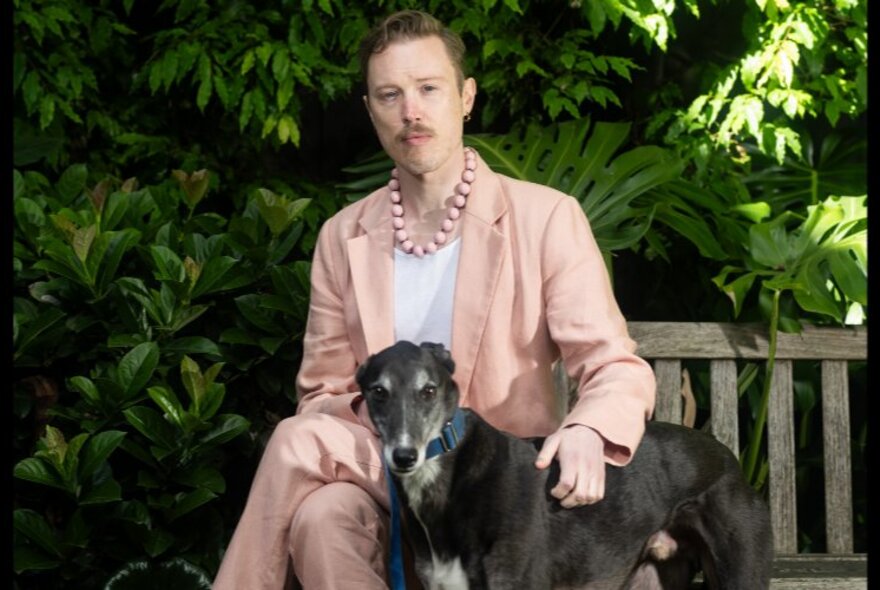 Author Patrick Lenton, seated on a wooden bench in front of a leafy green garden, wearing a pale pink suit and pink beaded necklace, patting a greyhound dog.