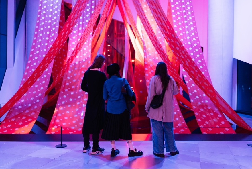 People inside a gallery space looking at an entrance with large red strips of fabric hanging from the ceiling. 