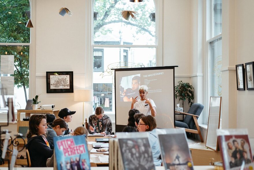 A classroom setting with kids seated at a table and watching their teacher who stands in front of a screen with a projection on it, large windows in the wall behind showing glimpses of trees and the street outside.