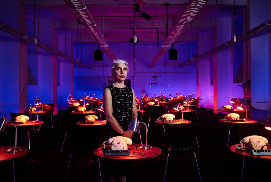 A person standing in a large room filled with small tables, each with a vintage phone and desk lamp, part of an interactive theatre installation at the Malthouse Theatre.
