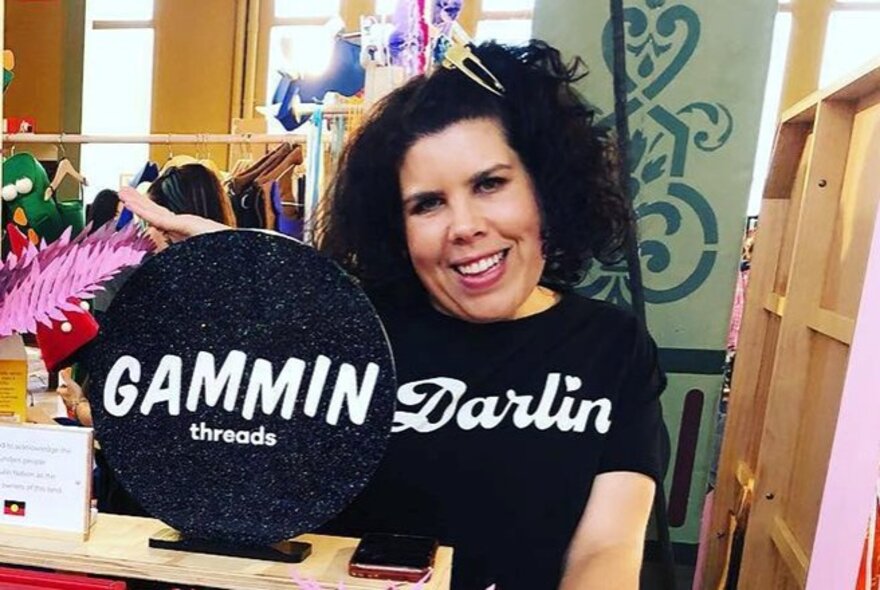 A woman with dark curly hair smiles at the camera while standing behind a table with a round black sign that reads GAMMIN threads. She is wearing a black t-shirt with 'Darlin' written in white font.