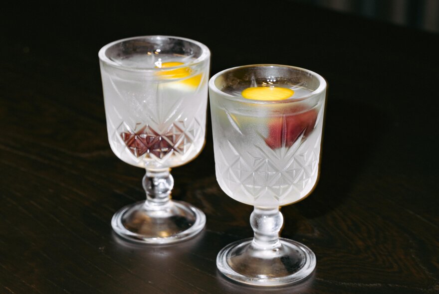 Two clear, chilled cocktails in short-stemmed crystal glasses, each with a slice of lemon and a plum as garnish.