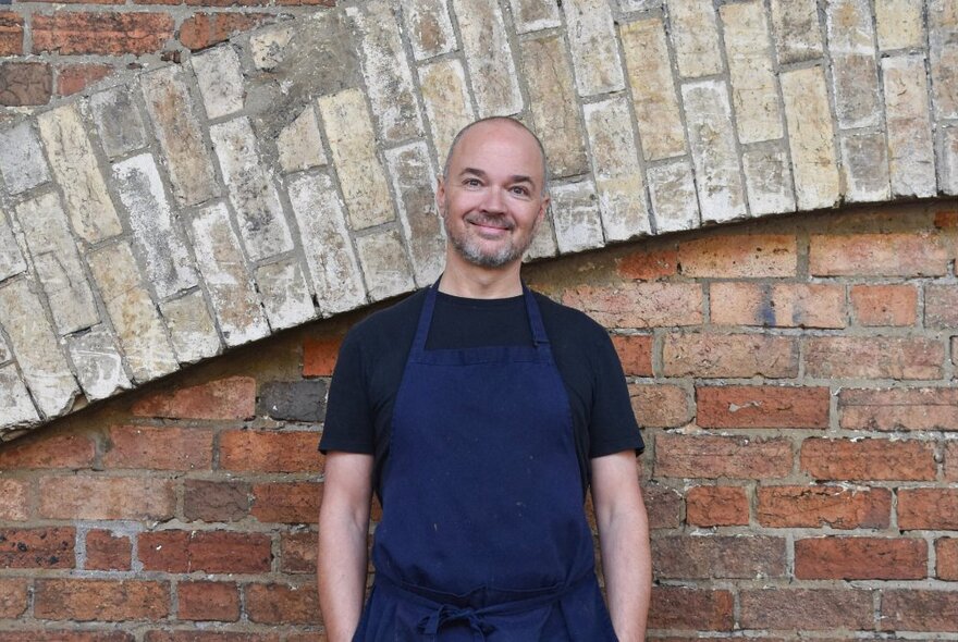Chef Tobie Puttock wearing an apron and smiling, standing in front of a brick wall.
