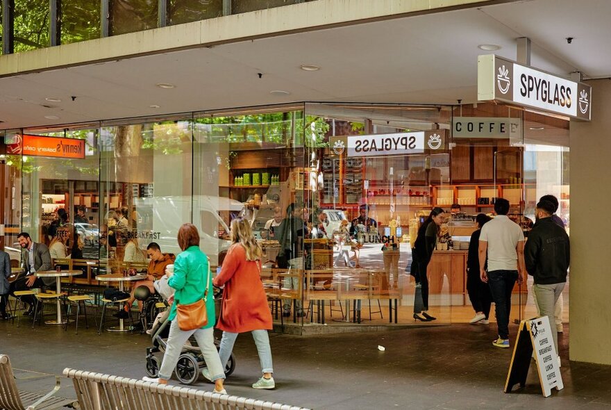 People walking past a coffee shop's glass storefront with indoor and outdoor tables.