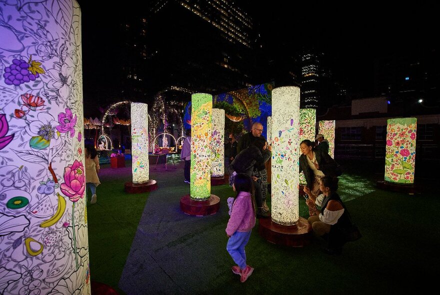 People passing illuminated pillars with floral artwork in the darkness.