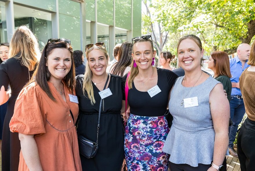 Four smiling women wearing nametags, posing for a photograph in front of a glass building.