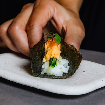 A gourmet hand roll with rice and spanner crab being held together by a hand while it rests on a small white plate.