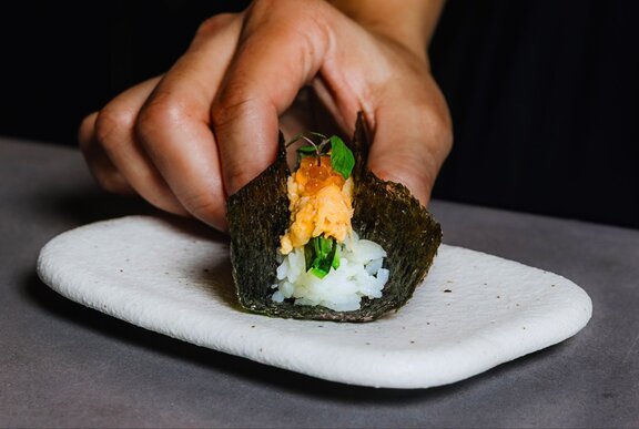 A gourmet hand roll with rice and spanner crab being held together by a hand while it rests on a small white plate.
