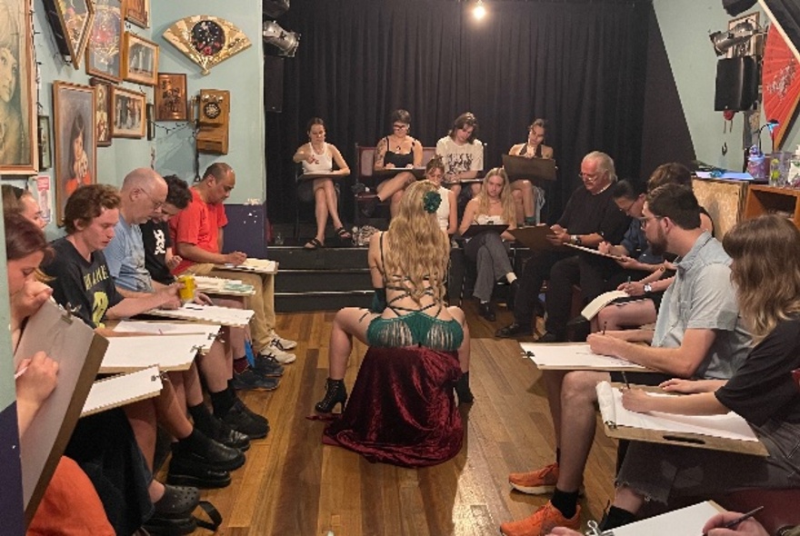 A crowded indoor life drawing session with a woman in burlesque costume as the subject with people arranged around her with large sheets of paper on their laps. 