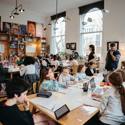 Kids seated around large tables writing and interacting with each other during a creative writing workshop, in a bright studio space.