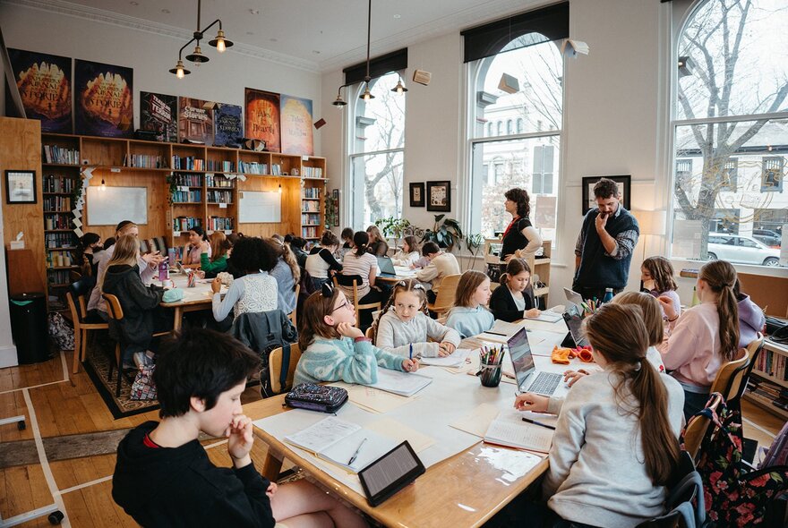 Kids seated around large tables writing and interacting with each other during a creative writing workshop, in a bright studio space.