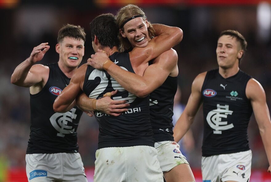 Four AFL players wearing Carlton tops congratulating each other in a hug on the football field.
