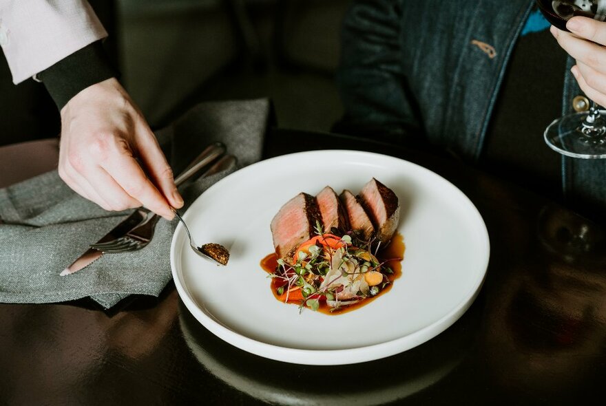 A hand placing a teaspoon of mustard on a white plate of slices of beef and a side of vegetables, at a restaurant table.