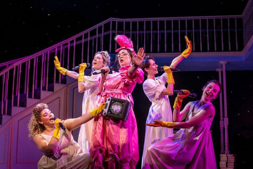 Female cast members from the musical Pride & Prejudice performing in Regency style costumes on a stage, all wearing yellow rubber gloves and singing into microphones they are holding.