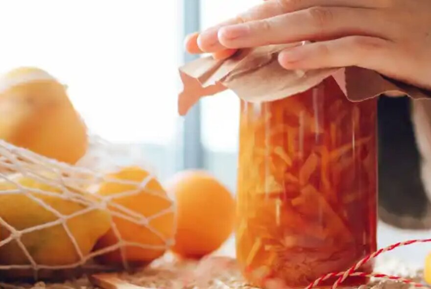 Hands pressing a piece of cloth over the top of a jar of marmalade, with a string bag of citrus fruit nearby.