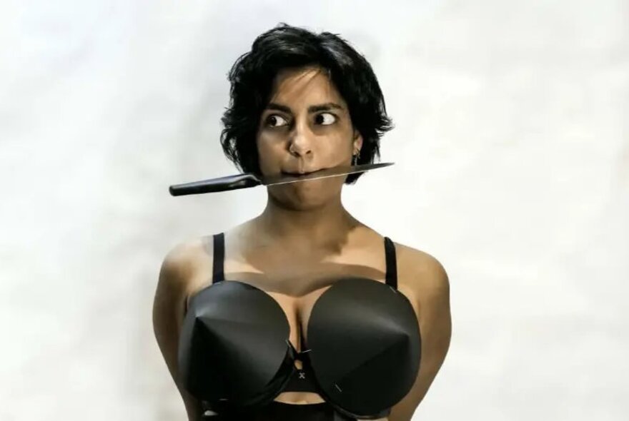 A person wearing a black brassiere with distinctive cone-shaped cups and biting onto a long, flat object in their mouth, against a white background.
