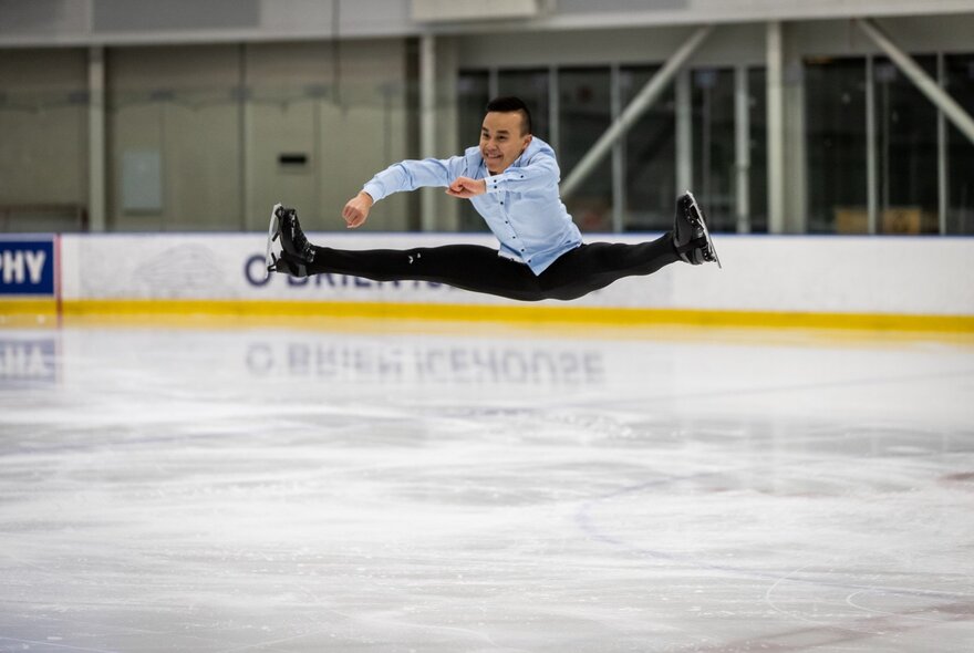 An ice skater performing an airborne split jump on an indoor ice rink.