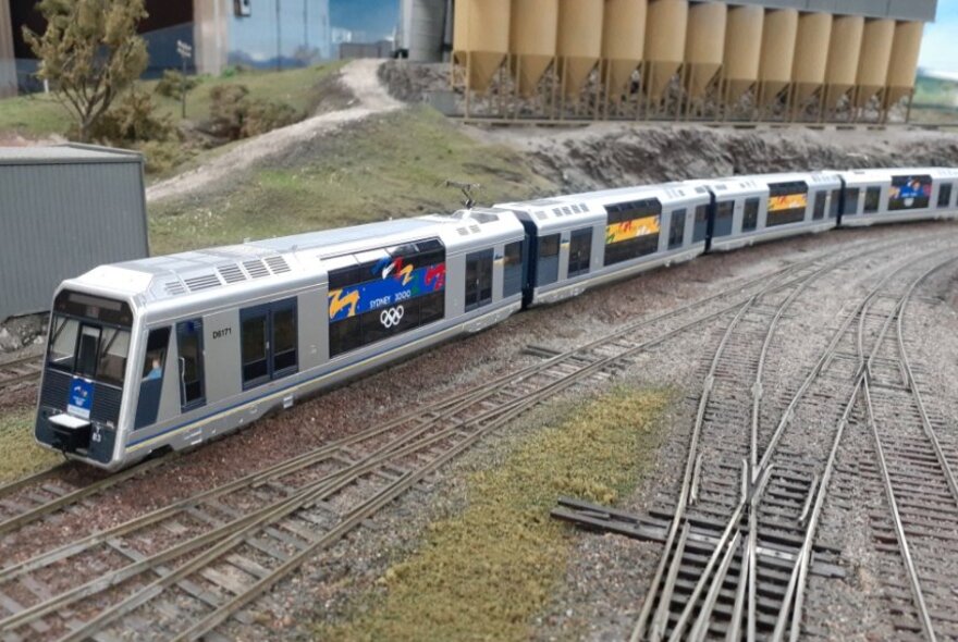 A modern-looking model passenger train on a model railway track, with a row of model silos behind it.