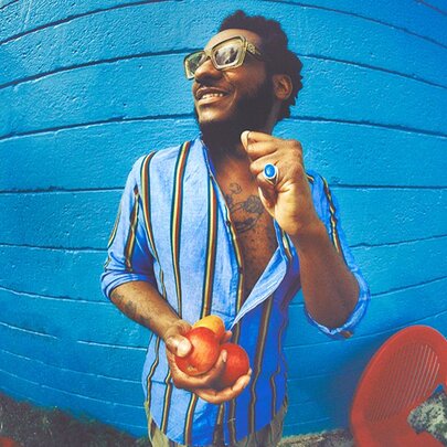 Leon Bridges, a man with dark hair, a beard, and glasses smiles while holding fresh peaches in front of a vibrant blue wall, photographed using a distorting fish eye lens.