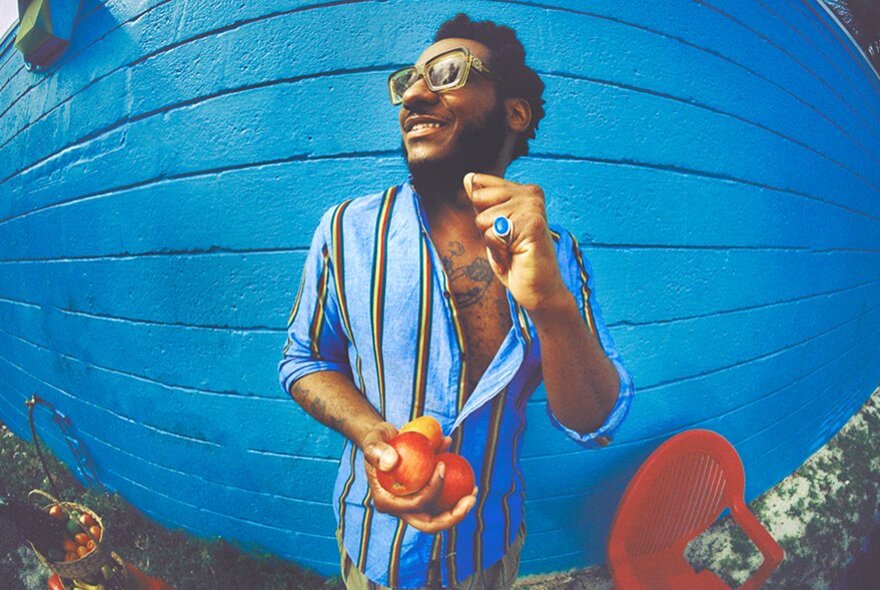 Leon Bridges, a man with dark hair, a beard, and glasses smiles while holding fresh peaches in front of a vibrant blue wall, photographed using a distorting fish eye lens.