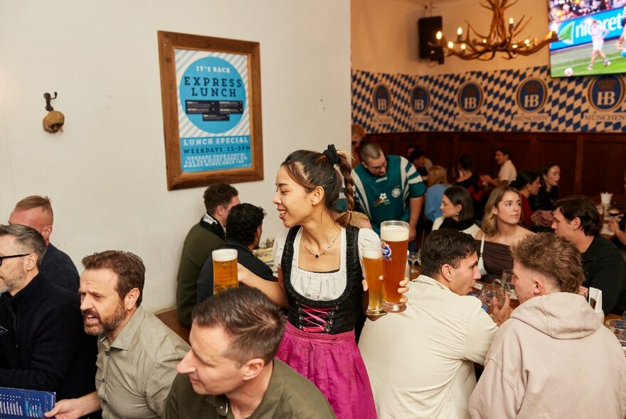 Bavarian-styled waitress wearing dirndl apron dress and carring steins of beer to busy tables with seated customers.