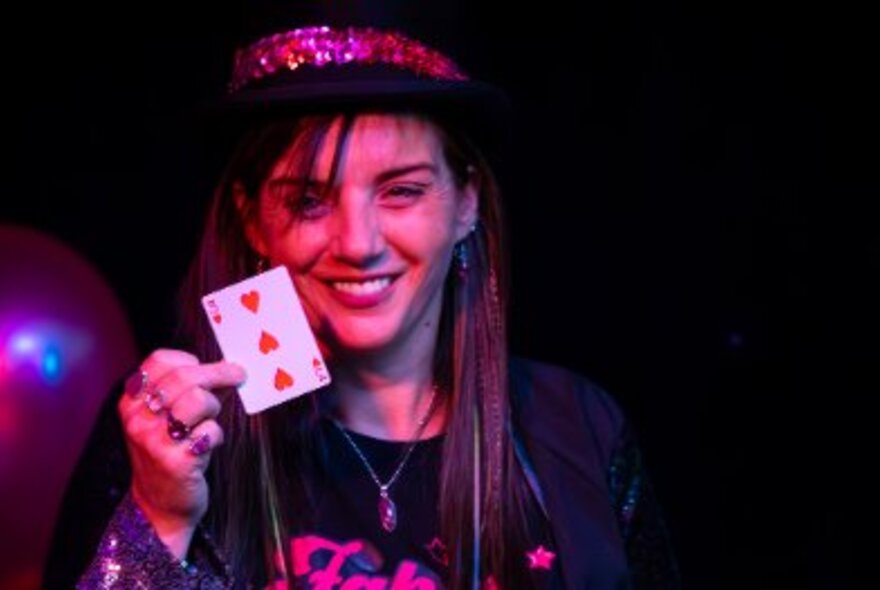 Woman wearing a black hat with pink ribbon performing on a stage, holding up a 3 of hearts playing card close to her face.
