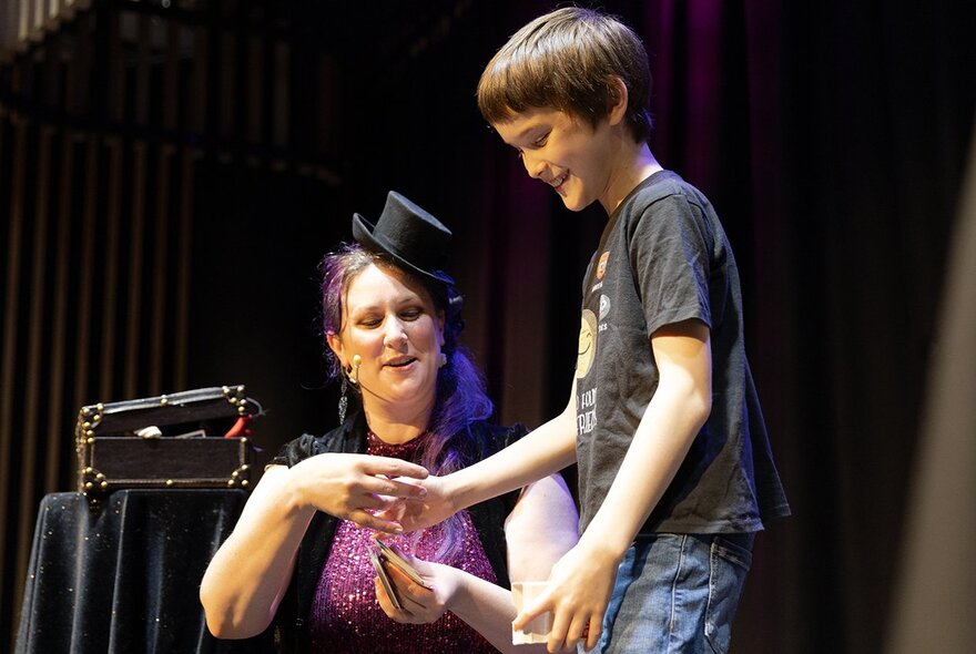 Woman wearing a top hat with case next to a young boy on stage.