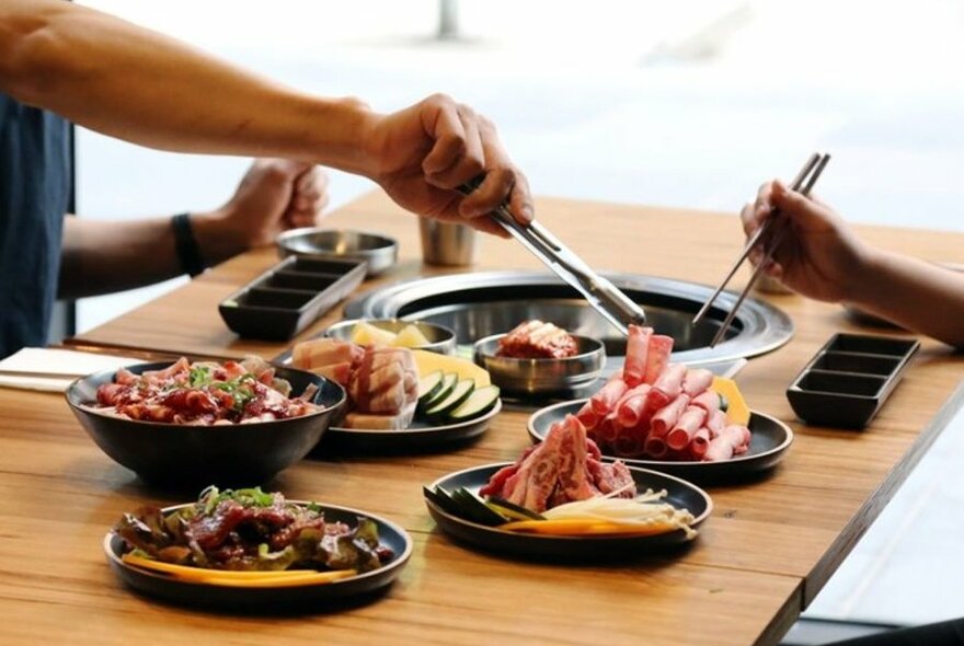 Two people with tongs picking meat up off a hot plate.