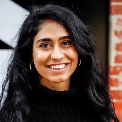 A woman with long black hair smiling openly in front of a brick wall with black and white lettering. 