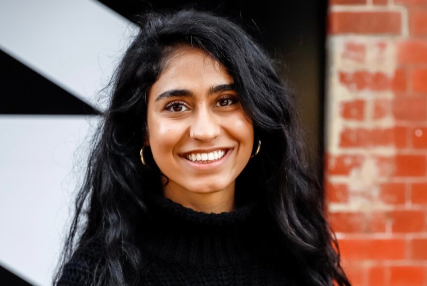 A woman with long black hair smiling openly in front of a brick wall with black and white lettering. 