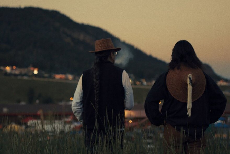 Film still depicting the back view of two people standing side by side looking out over twinkling village lights with a mountain in the background. 