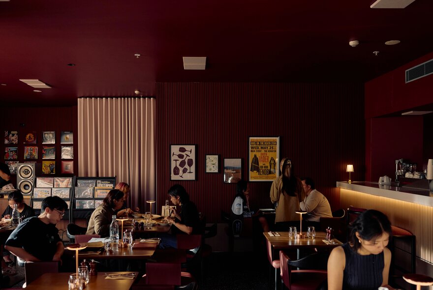 Burgundy walls and walnut-toned interior of Common Cuts restaurant, with patrons dining at tables, and a DJ vinyl record booth in the rear.