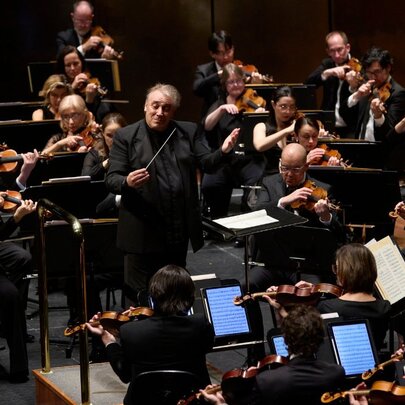 Black clad members of a symphony orchestra performing live on stage, in front of a conductor.