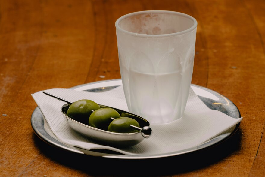 A frozen piccolo glass holding an ice-cold martini served alongside a small silver dish of green olives on a pick.