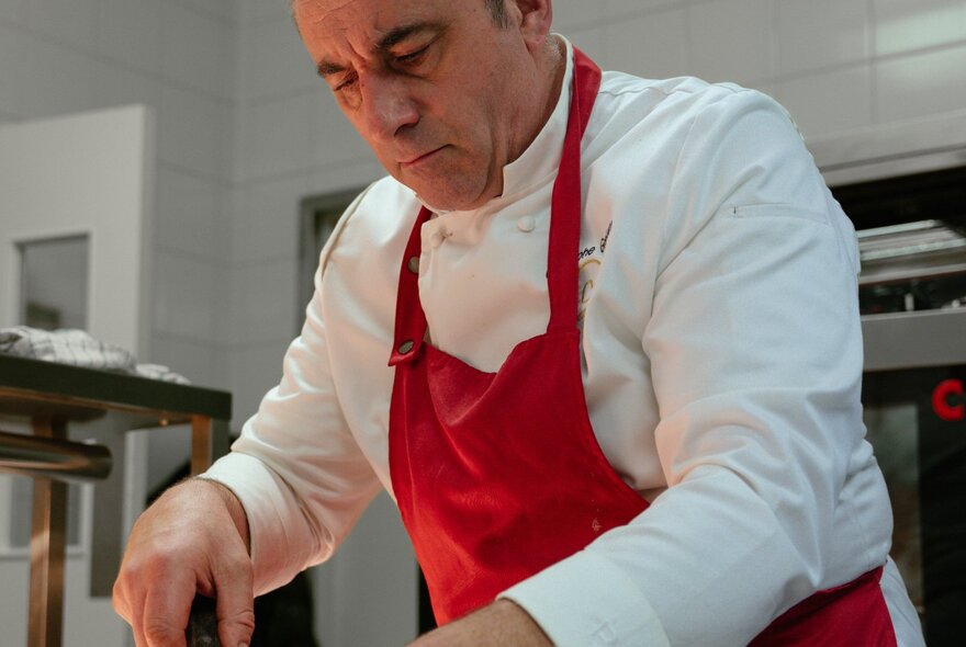 A chef in white wearing a red apron cooking in a commercial kitchen.