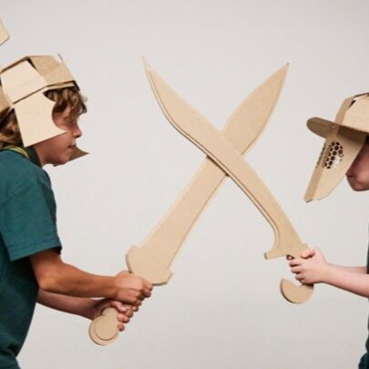 Two primary-school aged children, both wearing gladiator helmets and carrying swords constructed from cardboard, engaging in an interactive gladiator-themed creative play workshop.