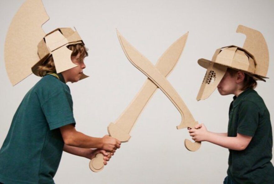 Two primary-school aged children, both wearing gladiator helmets and carrying swords constructed from cardboard, engaging in an interactive gladiator-themed creative play workshop.