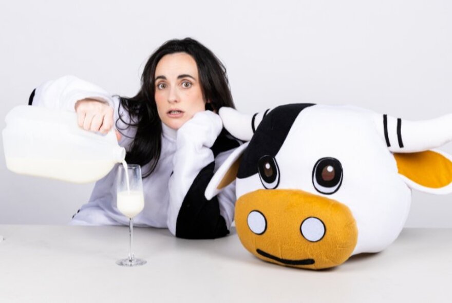 A woman pouring a glass on milk dressed in a cow suit, the head of the cow next to her on the table. 